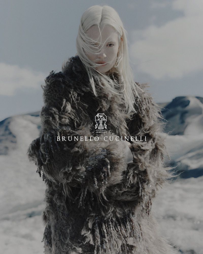 Model with long white hair in a textured grey fur coat against a snowy mountain backdrop with a cloudy sky.