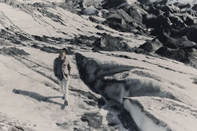A man walking across a vast glacier terrain wearing a tailored jacket and light trousers.