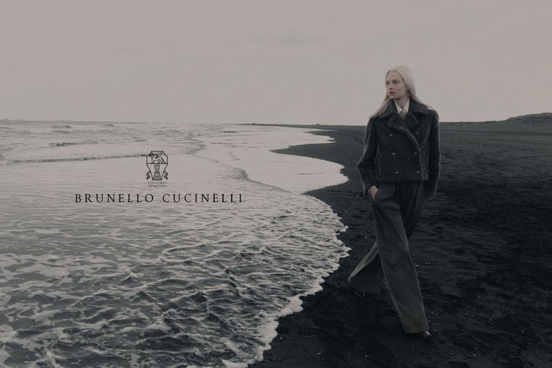 Model walking on a dark sandy beach wearing a textured coat and wide trousers under an overcast sky.