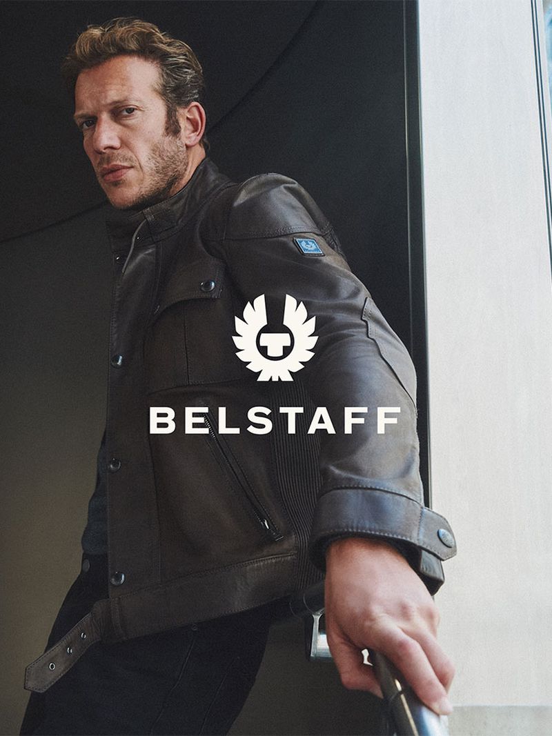 Man wearing a brown leather jacket and dark trousers standing outdoors near a building exterior.