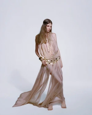Model in a sheer flowing dress wearing layered gold jewellery and headpiece against a plain studio background.