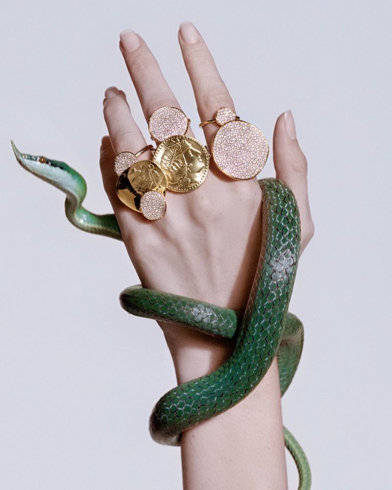 Close-up of a hand wearing multiple gold rings entwined with a green snake against a white background for the Ippolita Campaign.