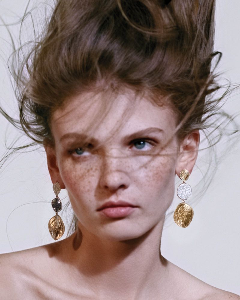 Close-up portrait of a woman with freckles and tousled hair wearing ornate dangling gold earrings against a light background
