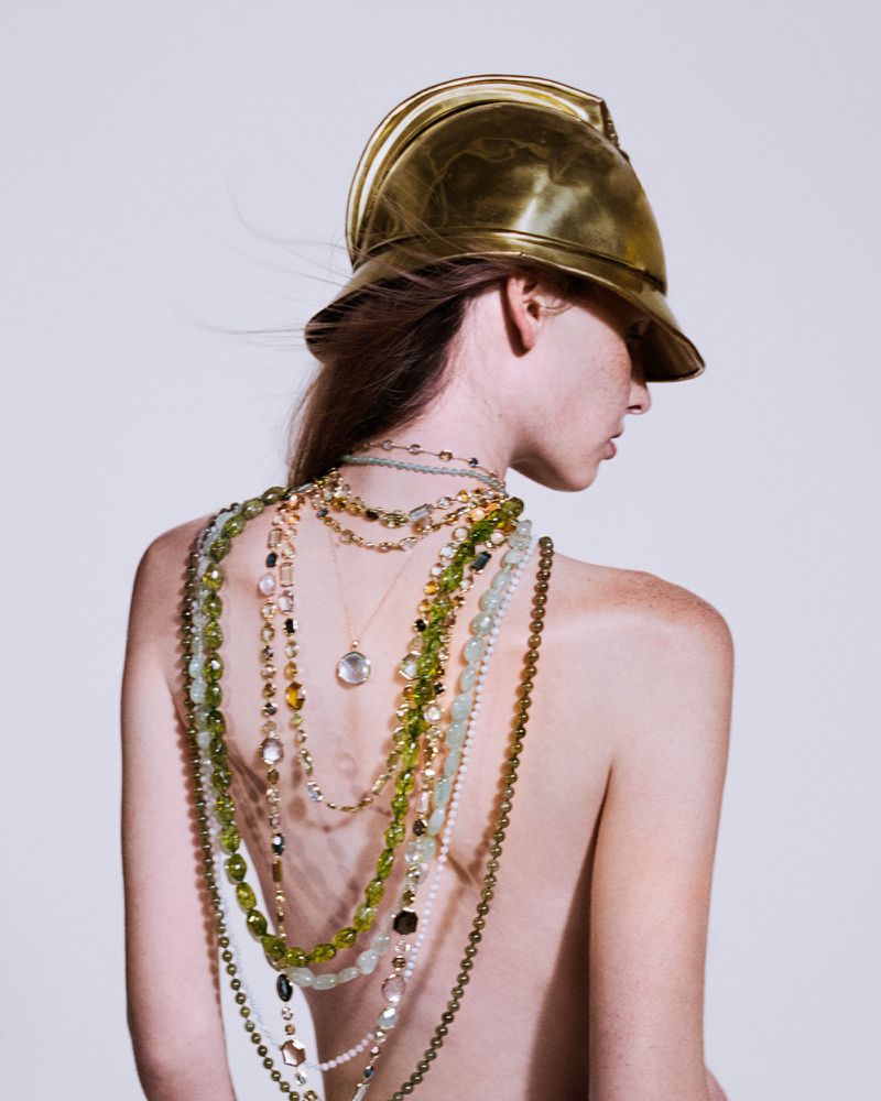 Model wearing a gold metallic helmet and layered gemstone necklaces draped down their back against a white background.