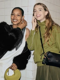 Two smiling models sharing white earbuds against a white tiled wall for Kate Spade Fall/Winter 2026 campaign.