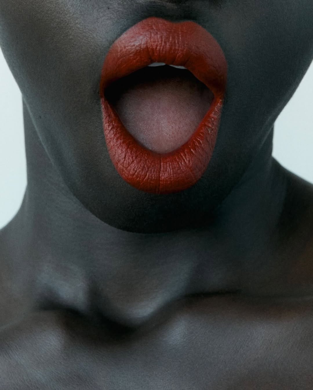 Close-up portrait of an African-American woman with dark skin and bold red lipstick, her mouth open, set against a clean, light background.