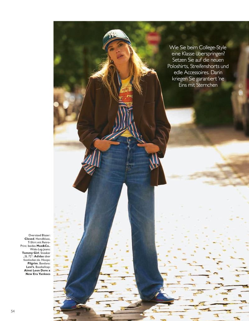 A woman in a baseball cap, blazer, striped shirt, t-shirt, and jeans stands on a brick road outdoors in a fashion editorial shot.