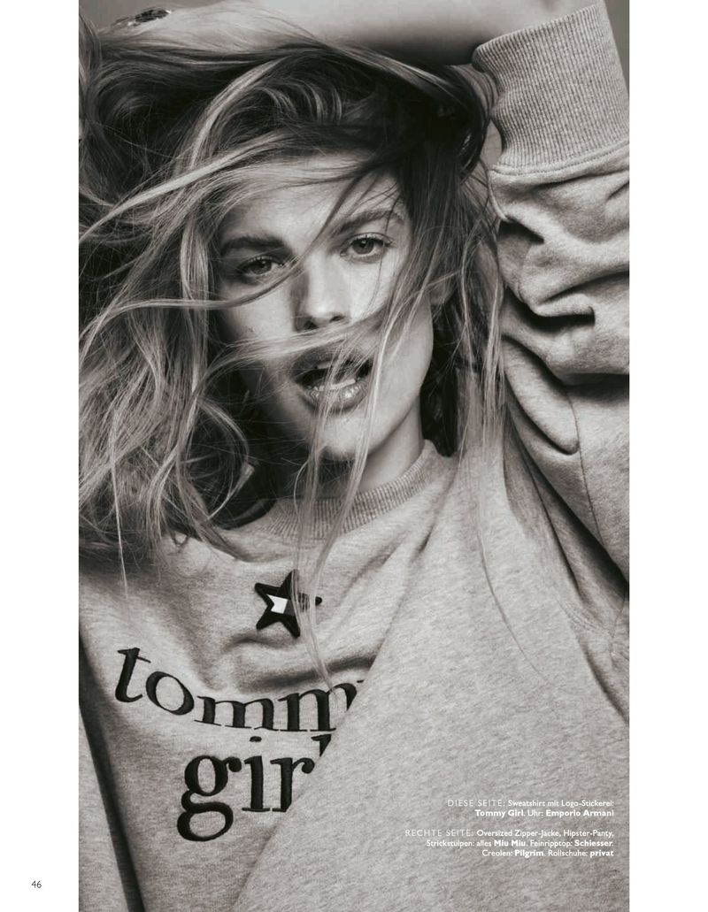 Blonde woman poses indoors wearing a grey Tommy Girl sweatshirt, photographed in black and white with soft lighting.