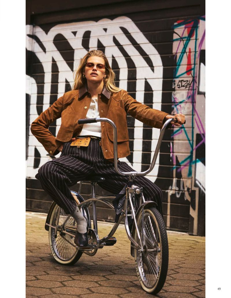 Woman in jacket and pinstripe pants poses on a vintage bicycle in an urban setting with graffiti wall backdrop, creating a fashionable outdoor shot.