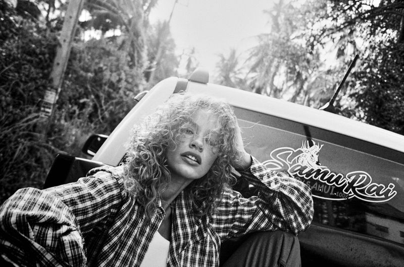 Monochrome fashion editorial image of Tess Carter leaning against a truck in a plaid shirt with tropical trees in the background.
