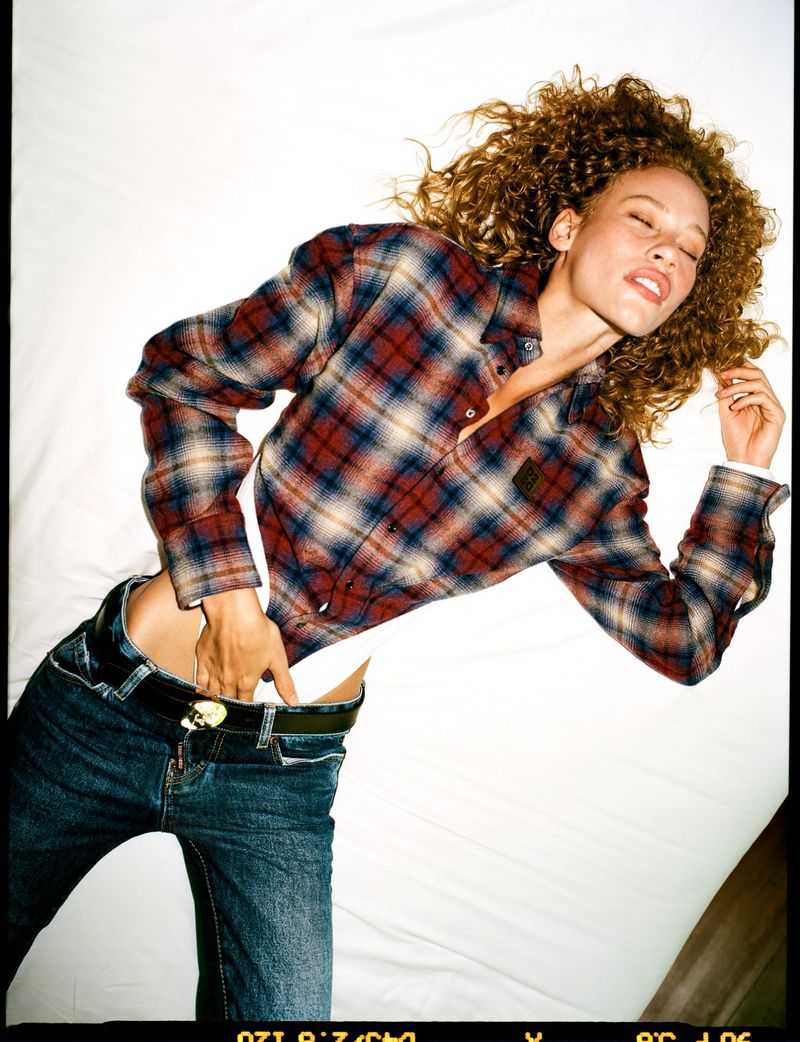 Model with curly hair wearing plaid shirt and blue jeans lying on a white background for Vogue Netherlands editorial