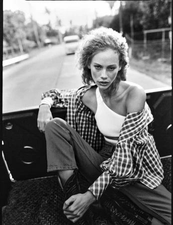 Model with curly hair wearing a plaid shirt and white crop top seated in the back of a vehicle on a road
