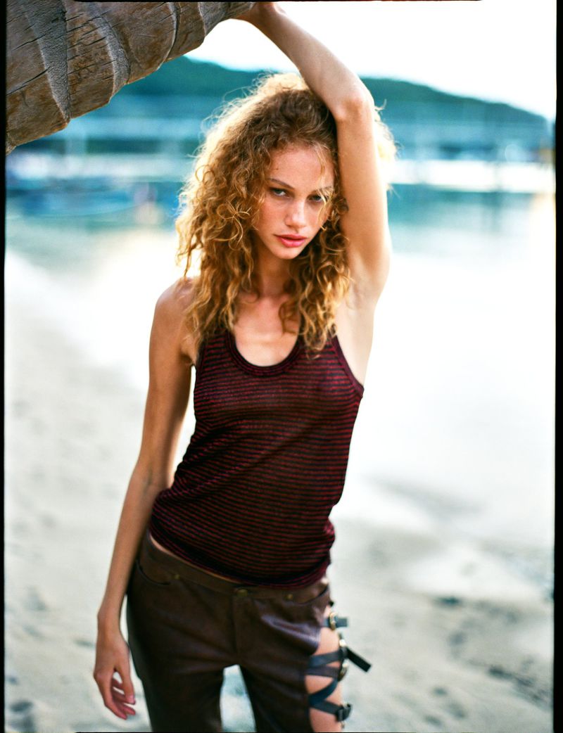 Model with curly hair in a dark striped tank top and trousers leaning against a palm tree on a sandy beach.