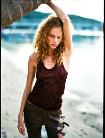 Model with curly hair in a dark striped tank top and trousers leaning against a palm tree on a sandy beach.