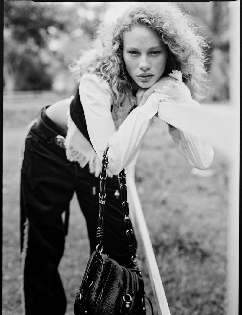 Model with curly hair leaning on a rail outdoors wearing a white blouse and dark trousers for Vogue Netherlands