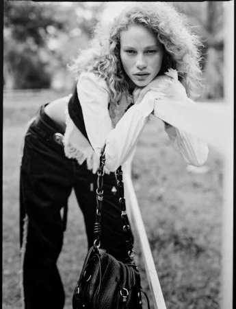 Model with curly hair leaning on a rail outdoors wearing a white blouse and dark trousers for Vogue Netherlands