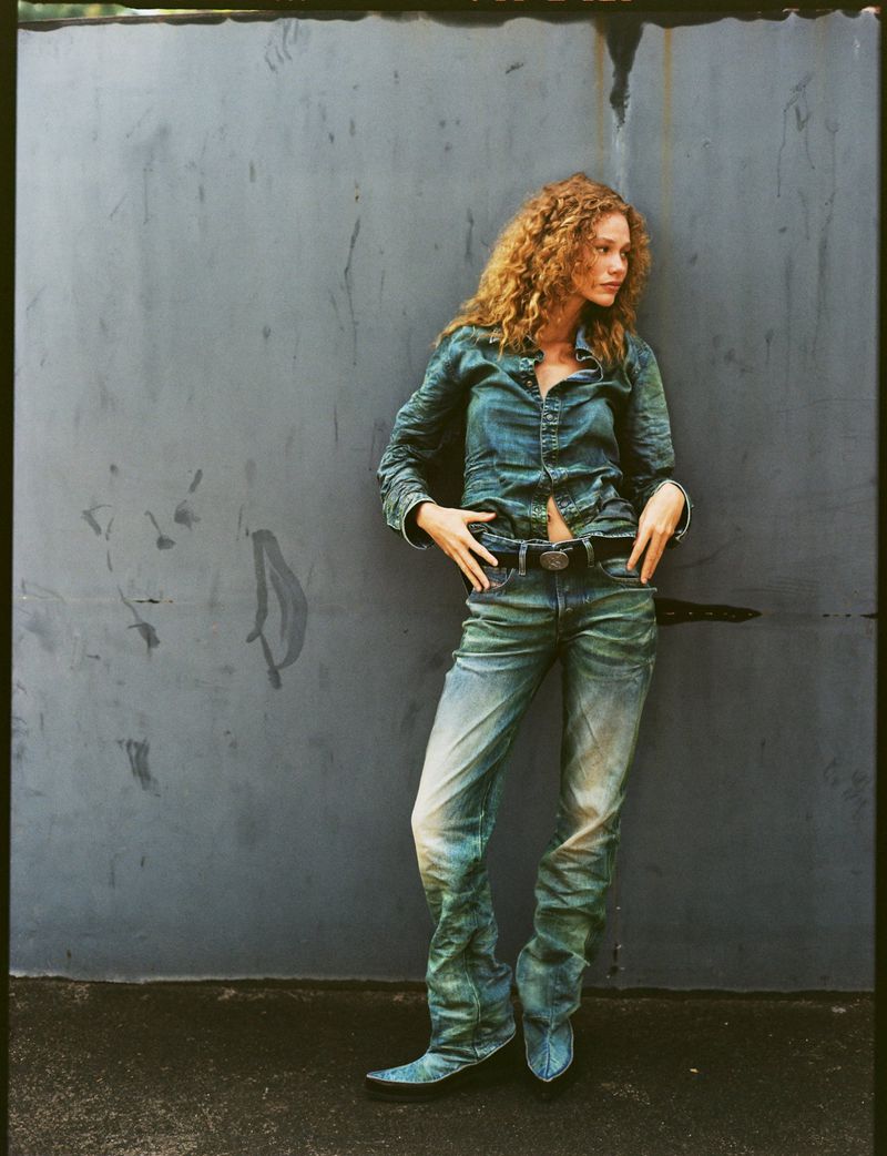 Model with curly hair leaning against a dark weathered wall wearing a blue denim shirt and matching jeans.