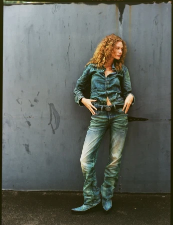 Model with curly hair leaning against a dark weathered wall wearing a blue denim shirt and matching jeans.
