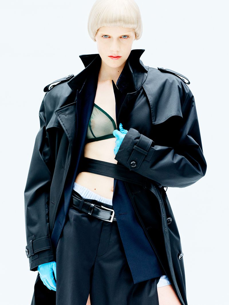 Model with blonde bowl cut wearing a black trench coat and blue gloves against a white background for D Repubblica