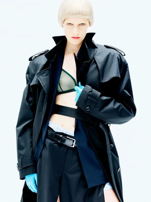 Model with blonde bowl cut wearing a black trench coat and blue gloves against a white background for D Repubblica