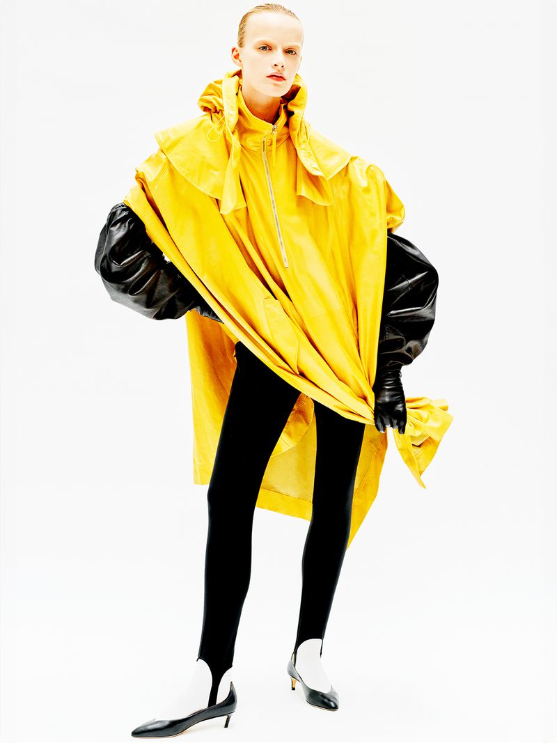Model wearing a bright yellow hooded jacket and black pants against a clean white seamless studio background.