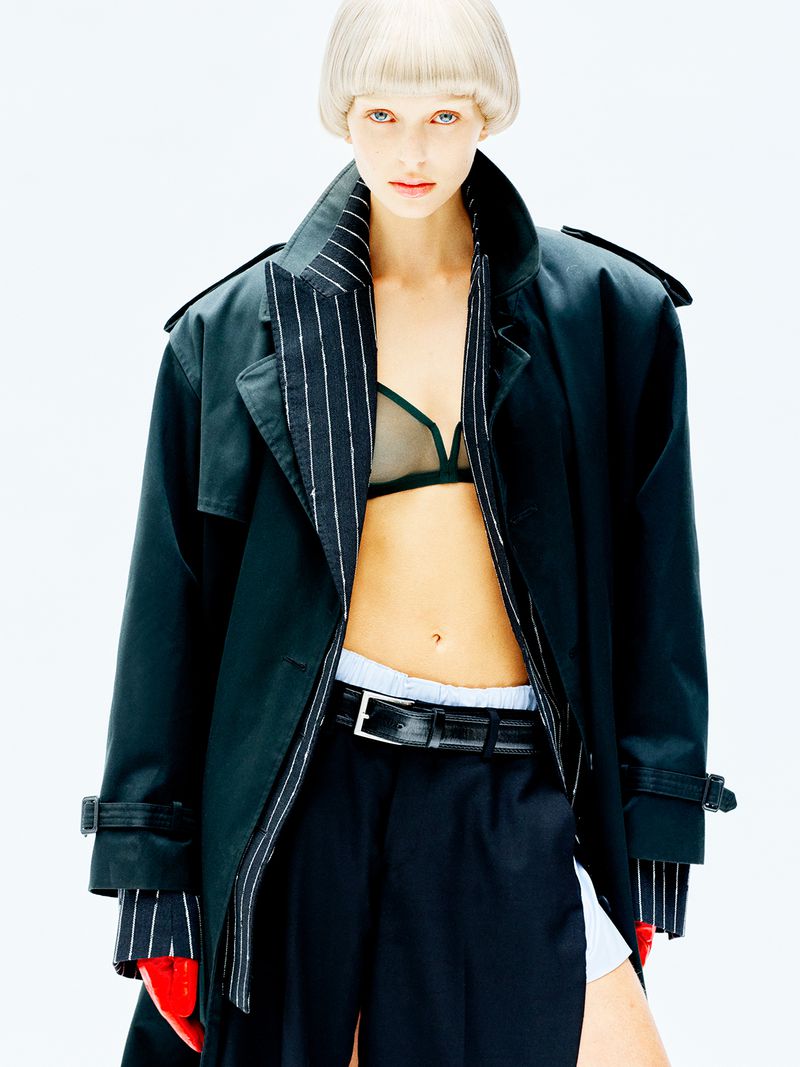 Model with blonde bowl cut hair wearing an oversized dark coat, sheer bra, and dark trousers against a white background.