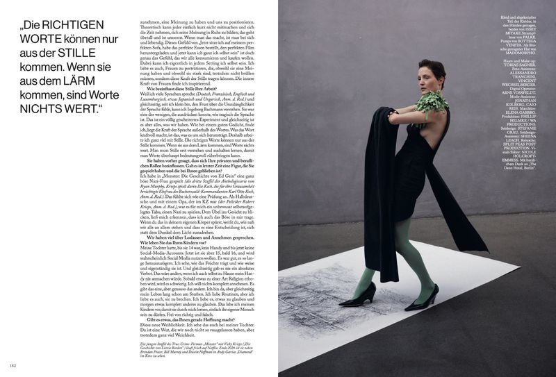 Vicky Krieps in a black dress and green tights standing on a printed paper sheet against a grey concrete wall