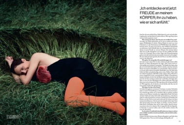 Vicky Krieps lying in tall grass wearing a dark dress and bright orange tights for Vogue Germany
