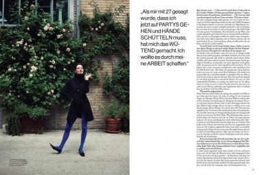 Vicky Krieps in a dark coat and blue tights standing beside a climbing rose bush for Vogue Germany ALLES ANDERS editorial