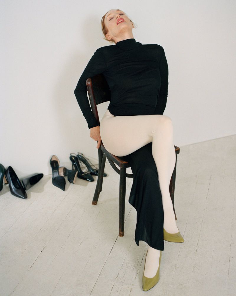 Julianne Moore seated on a dark wooden chair wearing a black turtleneck and white tights against a plain background.