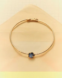 Thin gold bangle bracelet with a dark flower pendant resting inside a pale pink paper envelope on grid paper