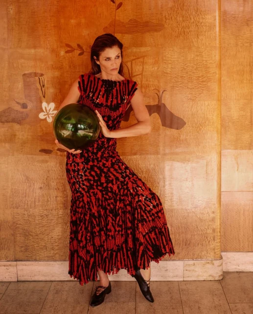 Helena Christensen holding a large glass sphere in a patterned red and black dress against a warm wooden wall