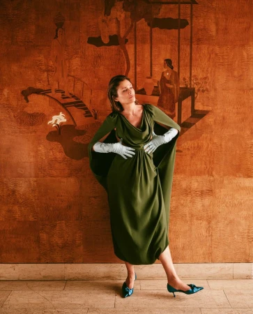 Helena Christensen in an olive green gown and light blue gloves standing against an ornate orange decorative wall