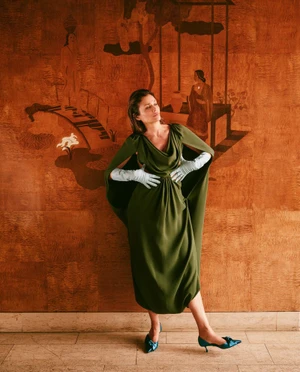 Helena Christensen in an olive green gown and light blue gloves standing against an ornate orange decorative wall