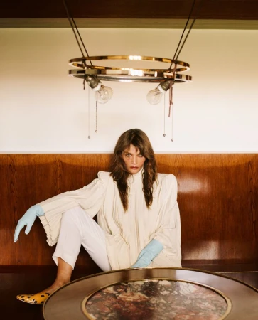 Helena Christensen seated in front of wood-paneled walls wearing a white blouse and light blue gloves