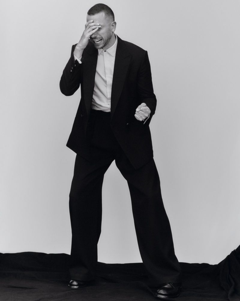 Man wearing a black suit and white shirt laughing with hand over face against a white studio background