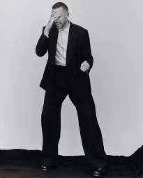 Man wearing a black suit and white shirt laughing with hand over face against a white studio background