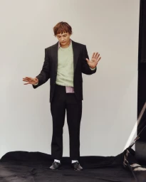 Man wearing a black suit jacket and trousers over a textured green knit sweater against a plain white studio backdrop.