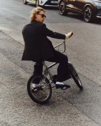 Man in a dark suit and sunglasses riding a bicycle on a paved street past parked cars