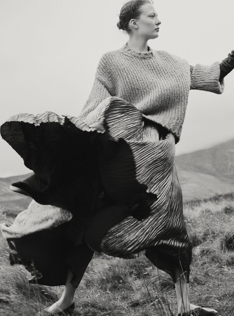 Model in a knit sweater and voluminous pleated skirt outdoors in a landscape photographed by Agata Pospieszynska for Harper's Bazaar UK