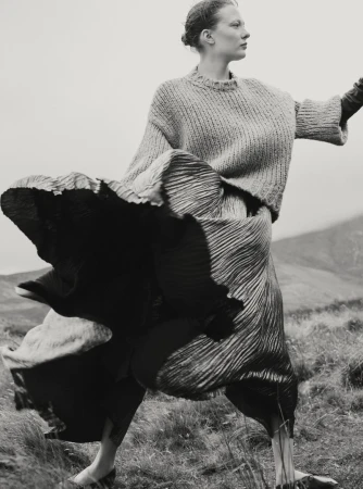 Model in a knit sweater and voluminous pleated skirt outdoors in a landscape photographed by Agata Pospieszynska for Harper's Bazaar UK