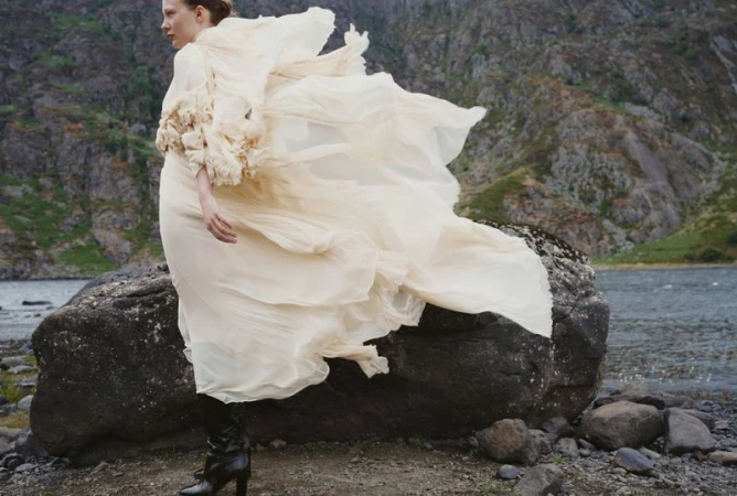 Model in flowing cream dress and dark boots standing on rocky shore against mountainous backdrop for Harper's Bazaar UK.