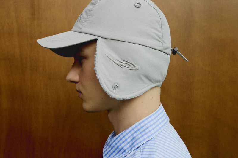 Profile portrait of a young man wearing a light grey earflap cap and a blue and white checkered shirt against a wooden background.