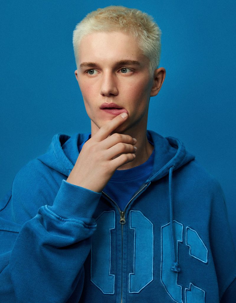 Young man with bleached blonde hair wearing a blue zip hoodie against a blue backdrop for Bershka