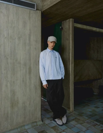 A male model leaning against a concrete pillar wearing a light blue button-down shirt, dark trousers, cap, and light shoes.