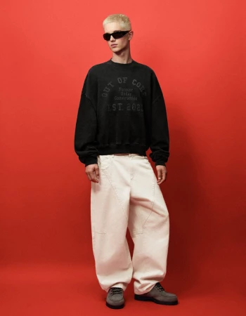 Young man with bleached hair wearing black sunglasses, a dark sweatshirt, and white wide-leg trousers against a solid red background