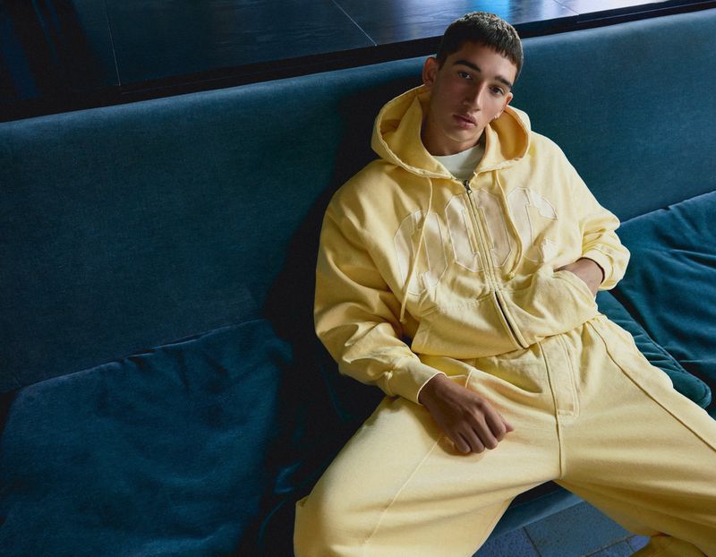 Young man wearing a yellow zip-up hoodie and matching trousers seated on a dark teal velvet couch.