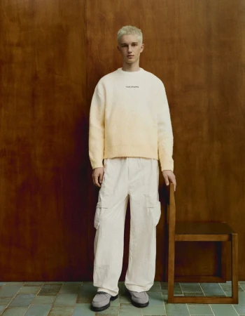 Young man with dyed blonde hair wearing a white sweater and light trousers standing beside a wooden chair.