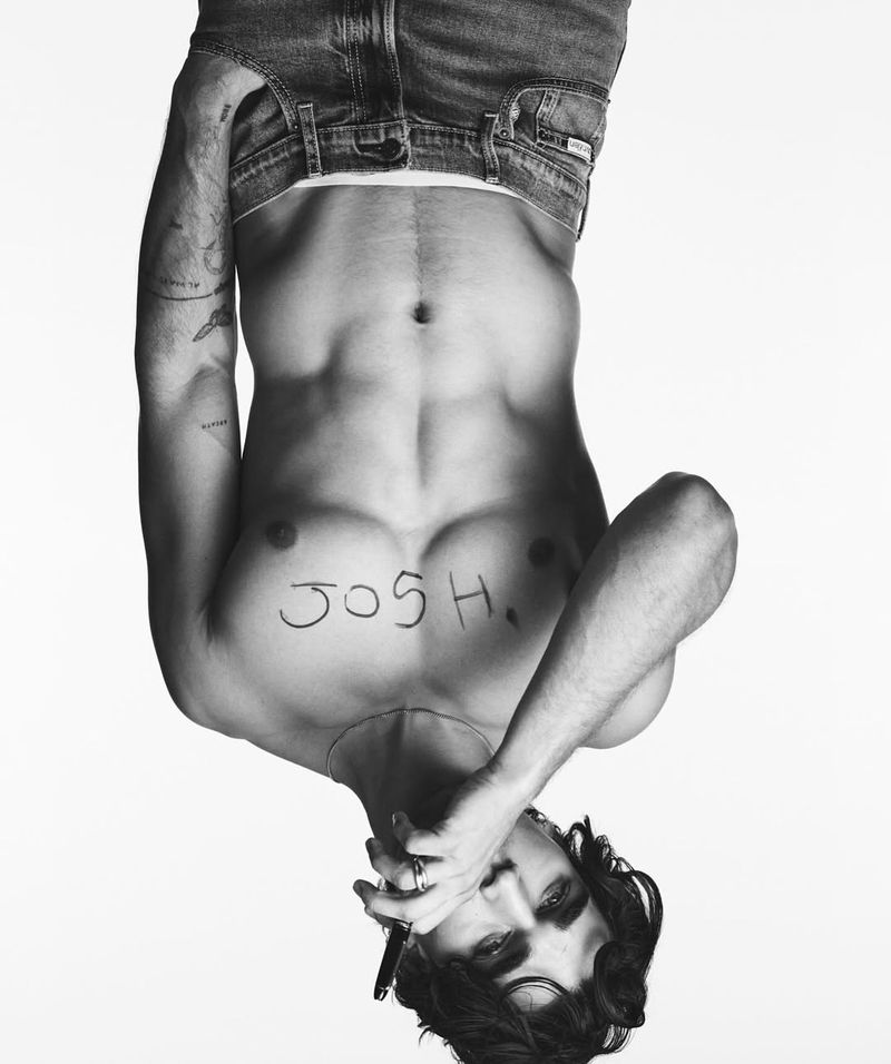 Black and white portrait of a shirtless adult male with abs and dark hair with jeans around his waist against a plain white background.