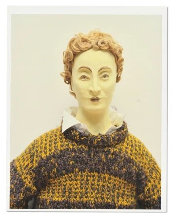 Portrait of a mannequin with short curly blonde hair wearing a striped knit sweater and white collared shirt.